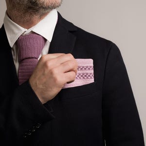 May include: A black blazer with a white collared shirt and a mauve knitted tie. A matching mauve pocket square with a geometric pattern is in the breast pocket. The image is a close-up of the upper body.
