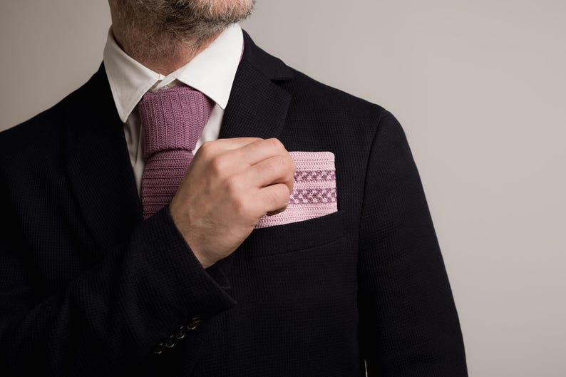 May include: A black blazer with a white collared shirt and a mauve knitted tie. A matching mauve pocket square with a geometric pattern is in the breast pocket. The image is a close-up of the upper body.