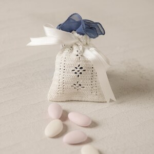 Può includere: Una borsa bianca in crochet con un nastro blu e un fiocco di raso blu. La borsa è piena di piccoli confetti di mandorle bianchi e rosa.