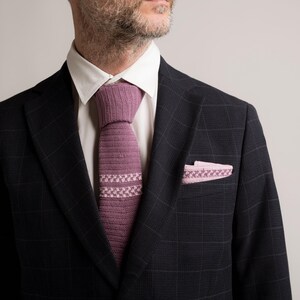 May include: A dark navy blue plaid suit jacket with a white dress shirt. The suit is accessorized with a knitted mauve tie and a matching pocket square. The tie and pocket square have a geometric design.