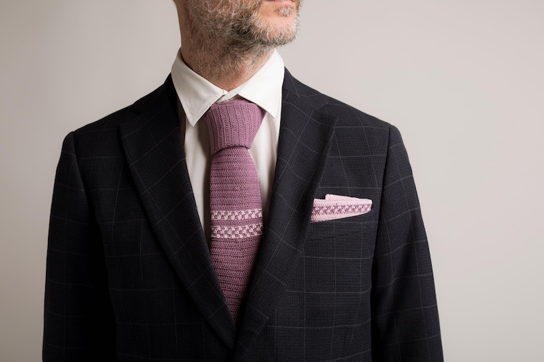 May include: A dark navy blue plaid suit jacket with a white dress shirt. The suit is accessorized with a knitted mauve tie and a matching pocket square. The tie and pocket square have a geometric design.