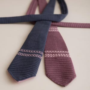 May include: Two knitted ties in navy blue and burgundy. Each tie features a decorative band of pink stitching near the tip. The ties are arranged in a crossed pattern against a neutral background, showcasing their texture and design.
