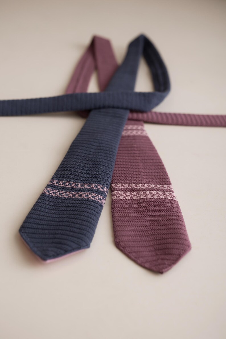 May include: Two knitted ties in navy blue and burgundy. Each tie features a decorative band of pink stitching near the tip. The ties are arranged in a crossed pattern against a neutral background, showcasing their texture and design.