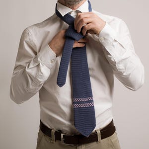May include: A navy blue tie with a textured pattern and a decorative band of pink and white squares. The tie is being knotted over a white dress shirt. The person is also wearing a brown belt and khaki pants.