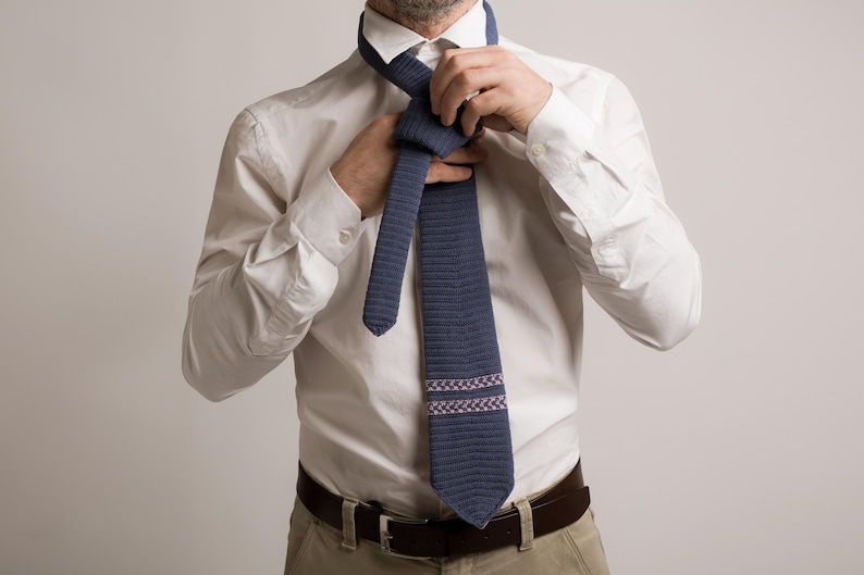 May include: A navy blue tie with a textured pattern and a decorative band of pink and white squares. The tie is being knotted over a white dress shirt. The person is also wearing a brown belt and khaki pants.