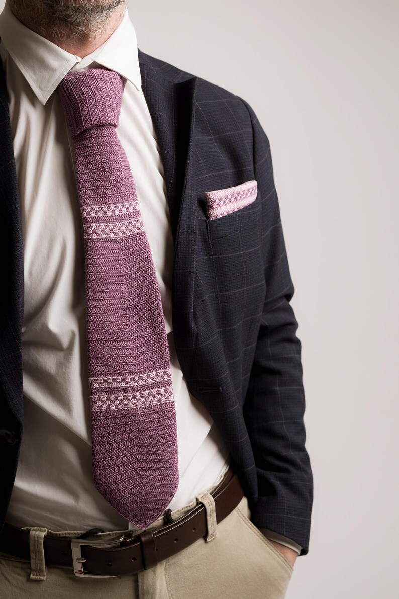 May include: A mauve knitted tie with a geometric pattern, worn with a white dress shirt and a navy blue plaid blazer. A matching pocket square is visible in the blazer's pocket. The tie and pocket square add a pop of color to the outfit.