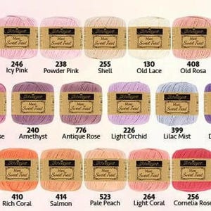 May include: A collection of colorful yarn skeins in various shades of pink, purple, orange, and red. Each skein has a label with a color name and number. The yarn is arranged in rows, showcasing a wide range of hues for crafting projects.