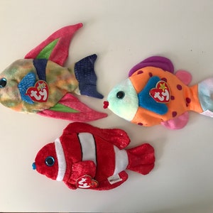 May include: Three colorful plush fish toys with Ty tags. The fish are a blue, green, and pink fish, an orange and pink fish, and a red and white fish.