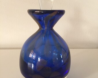 May include: A cobalt blue glass bottle with a clear glass stopper. The bottle has a white band around the bottom and a swirl of gold paint.