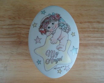 May include: A white porcelain trinket box with a round shape. The lid features a colorful illustration of an angel with a flower crown, sitting on a yellow star. The text "My Angel" is written on the star. The box is decorated with blue and pink stars.