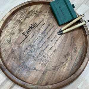 Könnte beinhalten: Ein hölzernes Farkle-Spieltablett mit geätzten Punktdetails und einem dekorativen Blätterrand. Enthält ein grünes Notizbuch und zwei Bambusstifte, ebenfalls mit der Aufschrift "Farkle". Das Tablett ist rund und aus hellbraunem Holz.