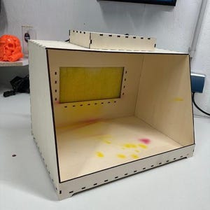 Può includere: Una cabina di verniciatura in legno chiaro con un'apertura rettangolare e un filtro. La cabina ha un piccolo pezzo superiore separato. Schizzi di vernice gialla, rosa e arancione sono visibili all'interno. La cabina è su una superficie bianca.