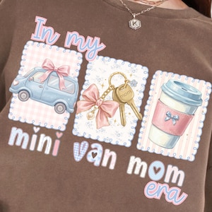 Könnte beinhalten: Braunes Sweatshirt mit dem Text "In my mini van mom era". Das Design zeigt einen hellblauen Van mit einer rosa Schleife, einen Schlüsselanhänger mit Schlüsseln und eine Kaffeetasse. Das Design hat ein rosa und blaues Farbschema.
