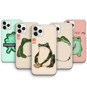 May include: A collection of phone cases featuring frog illustrations. Designs include a pixelated frog with "CENSORED" text, a green frog with a knife and "TEST ME" text, and other artistic frog depictions in various styles and colors.