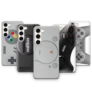 Könnte beinhalten: Eine Sammlung von Handyhüllen mit Retro-Videospielkonsolen-Designs. Die Hüllen zeigen ikonische Gamecontroller und Konsolenästhetik, darunter ein Super Nintendo, Sega Genesis, Sony PlayStation und andere. Die Hüllen sind in verschiedenen Farben erhältlich, darunter Grau, Schwarz und Weiß.