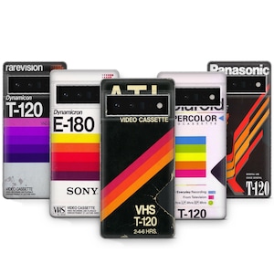 May include: Five different phone cases featuring a retro VHS tape design. The cases are black, white and red with colourful stripes. The text on the cases reads "T-120", "VHS", "Dynamicron", "Percolor", and "Panasonic".