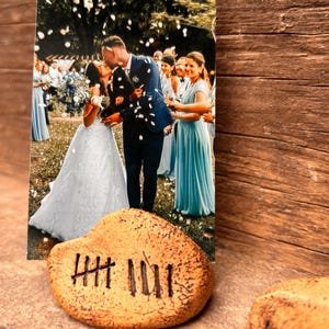 May include: A rectangular photograph of a wedding couple kissing, surrounded by guests in light blue attire, displayed in a tan, textured stone holder featuring hand-carved tally marks on the front surface, with a second stone visible nearby.