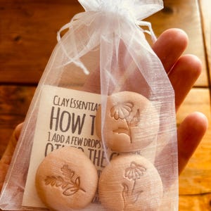 May include: A small white mesh bag tied with a ribbon, containing three round, light brown clay discs. Each disc features a different embossed floral design. A card inside the bag reads "Clay Essentials" and "How To".