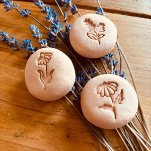 May include: Three round, light-coloured, floral-embossed stones rest on a wooden surface, accompanied by sprigs of blue lavender. Each stone features a different flower design. The arrangement creates a natural, rustic aesthetic.