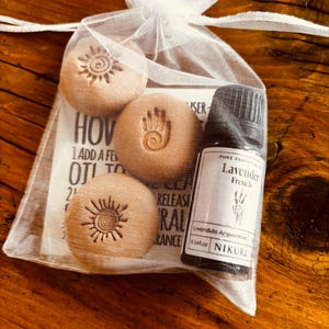 May include: A small white mesh bag containing three beige, round stones with sun and hand designs, and a small bottle of lavender essential oil. The oil bottle has a black cap and a white label with text.