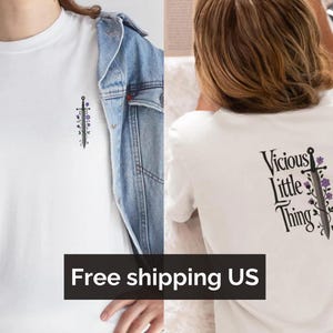 Może przedstawiać: Biała koszulka z małym wzorem miecza i kwiatów z przodu i tekstem "Vicious Little Thing" z wzorem miecza i kwiatów z tyłu.