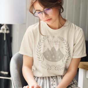 Op de afbeelding: Een vrouw draagt een wit T-shirt met een grijs grafisch ontwerp van een bergketen met sterren en de tekst "Velaris City of Starlights". Ze zit op een stoel en leest een boek.
