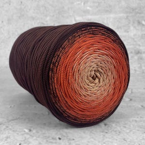 Op de afbeelding: Een bol garen met een kleurverloop, variërend van donkerbruin naar oranje en vervolgens naar een lichtbeige in het midden. Het garen is strak opgerold en geschikt voor brei- of haakprojecten. Het garen is waarschijnlijk van katoen.
