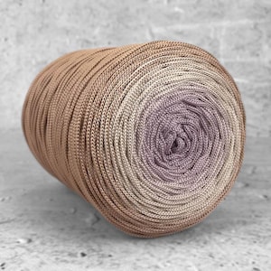 Op de afbeelding: Een grote, strak opgerolde bol garen met een kleurenschema. Het garen gaat over van lichtbruin naar een zachte lavendelkleur in het midden. De textuur van het garen lijkt glad en strak gesponnen, geschikt voor brei- of haakprojecten. Het garen wordt in grammen gemeten.
