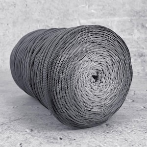 Op de afbeelding: Een strak opgerolde spoel grijs garen, rustend op een betonnen oppervlak. Het garen heeft een effen, neutrale grijze kleur, met een zichtbare geweven textuur. De spoel is cilindrisch, met het middengat zichtbaar.