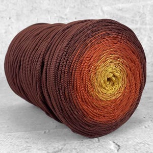 Könnte beinhalten: Eine zylindrische Spule aus geflochtener Kordel mit einem Farbverlauf, der von einem tiefen Dunkelbraun an den Außenkanten zu einem leuchtenden Orange und schließlich zu einem hellen Goldgelb in der Mitte der eng gewickelten Spule übergeht.