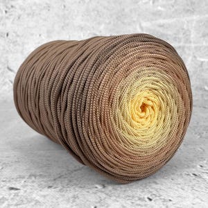 Può includere: Una bobina cilindrica di corda caratterizzata da una sfumatura di colore che passa da un marrone intenso sui bordi esterni a un giallo brillante al centro. La corda è avvolta strettamente in un motivo a spirale continua, creando una superficie.