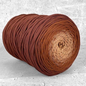 Può includere: Una grande bobina di filo marrone con un effetto sfumato, che passa da un marrone scuro a un beige più chiaro al centro. Il filo è avvolto strettamente, creando una superficie strutturata.