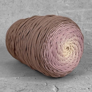 Peut inclure: Une pelote de laine aux tons marron, rose et crème. La laine est enroulée en spirale, avec des couleurs allant du marron au rose puis au crème au centre. La laine a un aspect texturé.