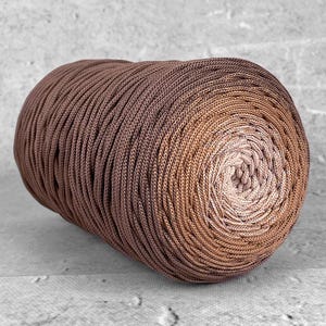 Op de afbeelding: Een strak opgerolde bol garen met een verloop van bruine tinten, variërend van donker chocoladebruin tot een licht beige centrum. De textuur van het garen lijkt strak geweven, wat duidt op een duurzaam materiaal dat geschikt is voor diverse knutselprojecten.