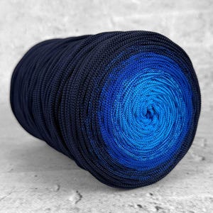 Può includere: Un grande gomitolo di filo con una sfumatura di blu scuro verso un blu brillante al centro. Il filo è ben avvolto e sembra essere fatto di una fibra liscia, forse sintetica. È appoggiato su una superficie grigio chiaro.