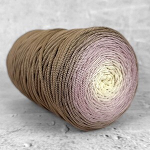 Puede incluir: Una gran madeja de hilo, bien enrollada, con un degradado de color. El hilo pasa de marrón oscuro a morado claro, luego a amarillo pálido en el centro. Adecuado para proyectos de tejido o ganchillo. El hilo es probablemente de algodón o fibra similar.