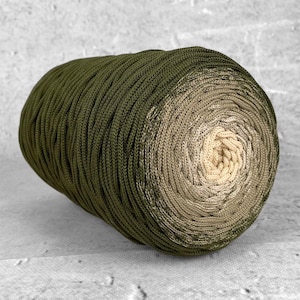 Puede incluir: Una gran madeja de hilo cilíndrica con un esquema de color degradado. El hilo pasa de un verde oliva oscuro a un beige claro en el centro. La textura del hilo parece estar tejida apretadamente.