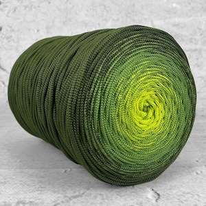 Puede incluir: Una bobina cilíndrica de cordón trenzado con un degradado de color que transita desde un verde oliva oscuro en los bordes exteriores hasta un amarillo brillante y vibrante en el centro. El cordón tiene una textura suave y tejida.