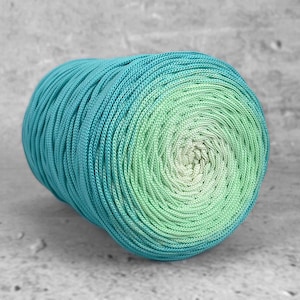 Puede incluir: Una bola de hilo bien enrollada en tonos turquesa y verde. El hilo pasa de un turquesa intenso en el exterior a un verde claro en el centro. El hilo se utiliza probablemente para proyectos de punto o ganchillo.