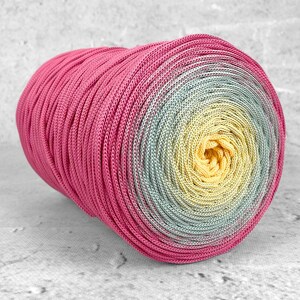 Op de afbeelding: Een cilindrische spoel van gevlochten koord met een kleurverloop dat overgaat van felroze aan de buitenranden naar zacht saliegroen, eindigend in een heldergeel midden. Het koord heeft een gladde en consistente textuur over het gehele oppervlak.