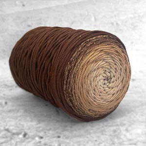 Peut inclure: Pelote de fil marron, présentant un dégradé de couleurs allant du brun foncé au beige clair. Le fil est étroitement enroulé, créant une surface texturée. Idéal pour les projets de tricot et de crochet.