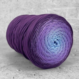 Puede incluir: Una gran madeja de hilo en tonos morados y azules. El hilo está bien enrollado, creando un efecto degradado del morado oscuro a un centro azul claro. La textura del hilo es visible, lo que sugiere un material suave y tejido.