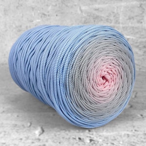 Può includere: Un grande gomitolo di filato con una sfumatura di colore. Lo strato esterno è azzurro, che sfuma nel grigio e infine in un rosa tenue al centro. Il filato è ben filato, creando una superficie strutturata.