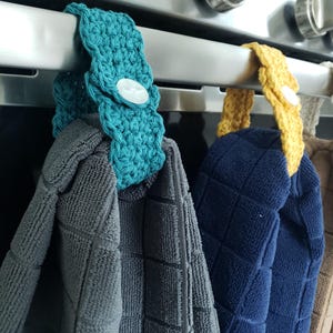 Pode incluir: Tr&ecirc;s porta-toalhas de croch&ecirc; pendurados em um rack de forno prateado. Os porta-toalhas s&atilde;o turquesa, amarelo e branco. As toalhas s&atilde;o cinza, azul e marrom.