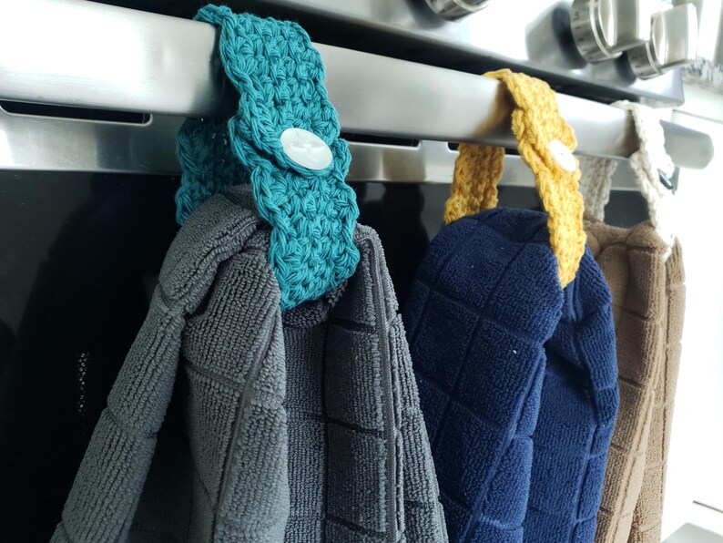 Pode incluir: Tr&ecirc;s porta-toalhas de croch&ecirc; pendurados em um rack de forno prateado. Os porta-toalhas s&atilde;o turquesa, amarelo e branco. As toalhas s&atilde;o cinza, azul e marrom.