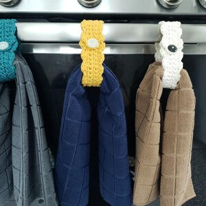 Pode incluir: Tr&ecirc;s porta-toalhas de croch&ecirc; em diferentes cores, turquesa, amarelo e branco, est&atilde;o pendurados em uma grelha de forno prateada. Cada porta-toalhas tem um bot&atilde;o no centro. Os porta-toalhas seguram toalhas cinzas, azuis e marrons.