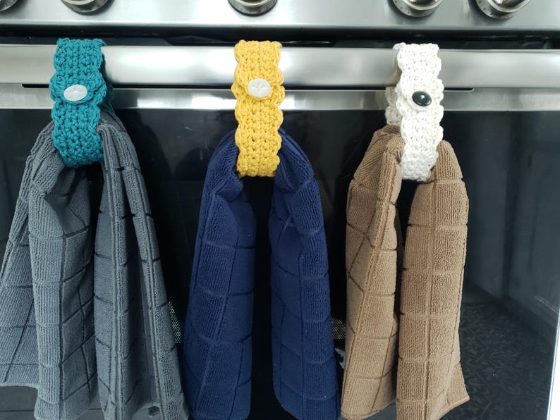 Pode incluir: Tr&ecirc;s porta-toalhas de croch&ecirc; em diferentes cores, turquesa, amarelo e branco, est&atilde;o pendurados em uma grelha de forno prateada. Cada porta-toalhas tem um bot&atilde;o no centro. Os porta-toalhas seguram toalhas cinzas, azuis e marrons.