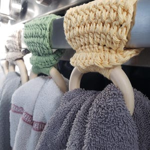 May include: Four crocheted towel rings in green, yellow, and beige hang on a silver oven rack. The rings are made of yarn and have wooden dowels. Two towels are hanging on the rings, one gray and one white with pink stripes.