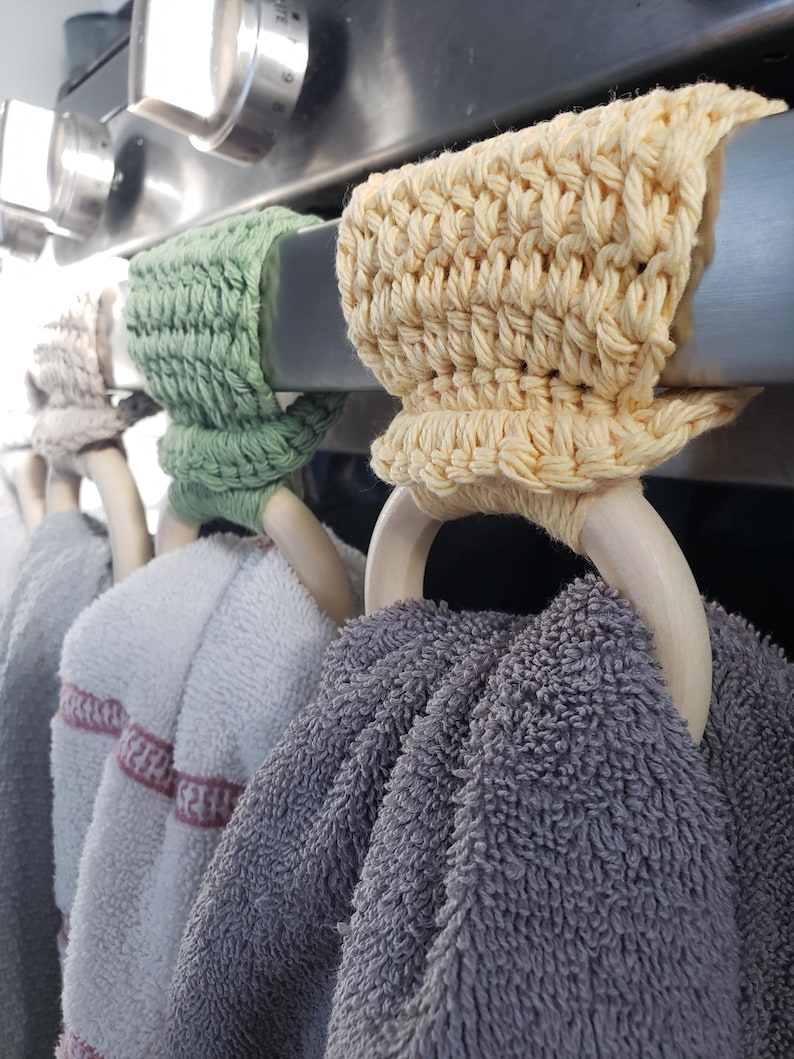 May include: Four crocheted towel rings in green, yellow, and beige hang on a silver oven rack. The rings are made of yarn and have wooden dowels. Two towels are hanging on the rings, one gray and one white with pink stripes.