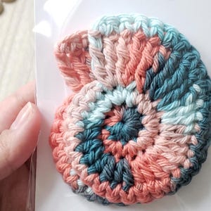 May include: A crocheted seashell in shades of pink, teal, and white. The seashell is a spiral shape and is made with a textured stitch.
